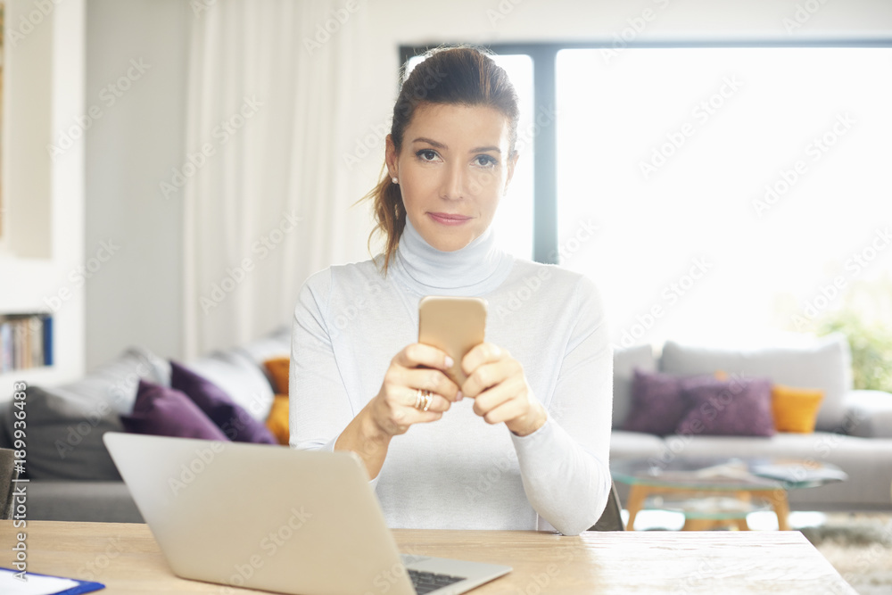 Home office. Confident woman talking on her mobile phone and using laptop while sitting at desk and working from home. 
