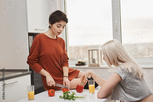 Indoor portrait of attractive hot woman sitting on table cutting tomotoe while her girlfriend drinks juice in kitchen. Lovely lgbt couple preparing healthy breakfast together. photo