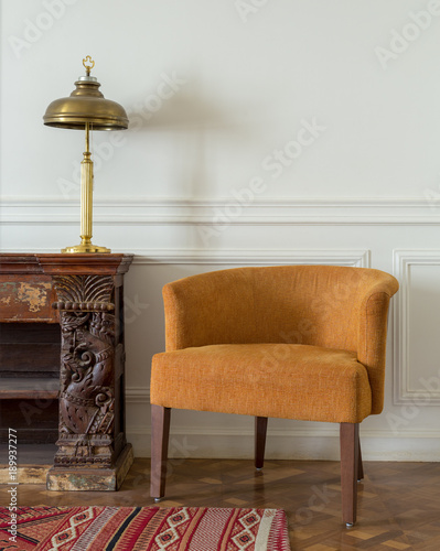 Interior shot of orange armchair and wooden vintage ornate sideboard and table lamp on off white wall and ornate carpet over wooden parquet floor photo