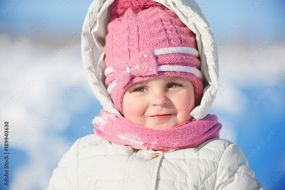 Close up portrait of adorable female child in warm winter clothing poses against snow background, enjoys fresh air and beautiful sunny frosty day, plays with children, happy to be outdoor