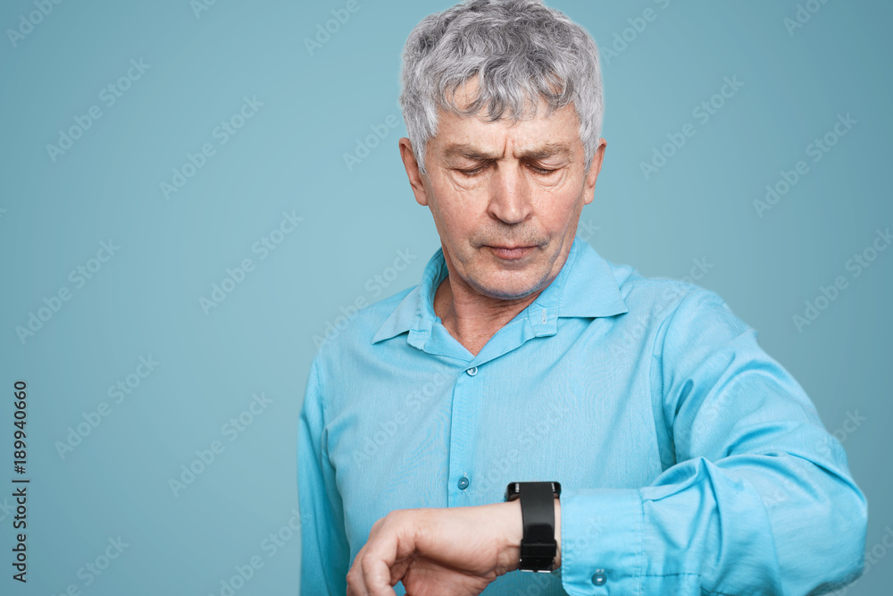 Senior male with serious expression looks at smartwatch with health application, controls his state, counts calories and measures blood pressure, isolated over blue background. Healthcare concept Senior male with serious expression looks at smartwatch with health application, controls his state, counts calories and measures blood pressure, isolated over blue background. Healthcare concept
