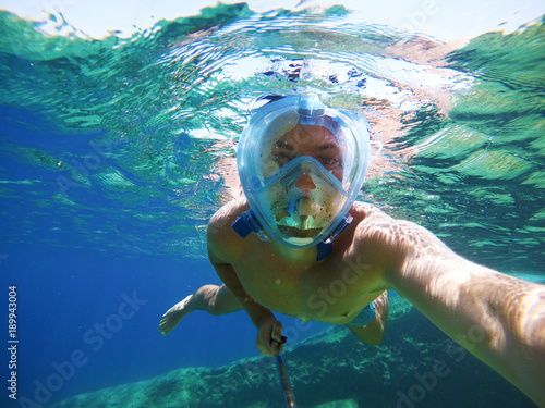 Young joyful handsome man taking a photo with a selfie stick while diving under the sea surface.