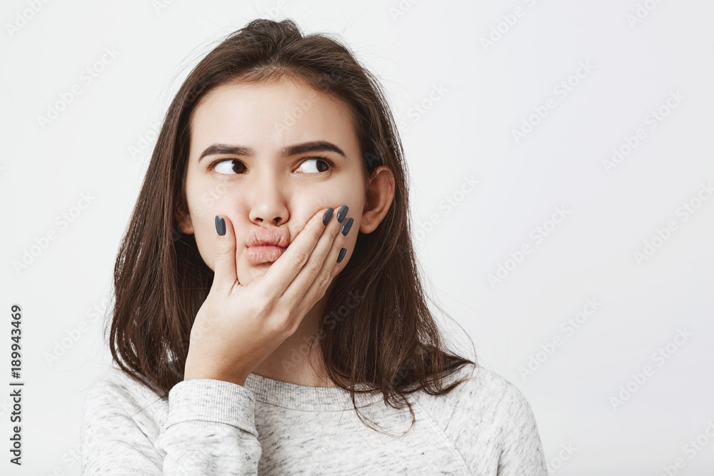 Close-up portrait of young female brunette with curious expression squeezing her face and thinking about something, isolated over white background. Girl makes up solution of how to avoid punishment