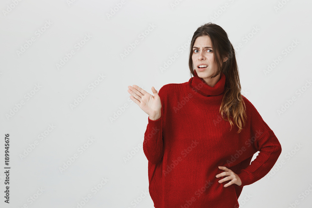 Waist-up portrait of young woman in red loose sweater with dark hair, frowning her face, standing against gray wall with copy space, showing opened palm, expressing her dissatisfaction and dislike Waist-up portrait of young woman in red loose sweater with dark hair, frowning her face, standing against gray wall with copy space, showing opened palm, expressing her dissatisfaction and dislike