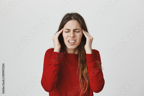 Stressed frustrated young female dressed in red sweater with closed eyes, frowns face, holds hands on temples, suffers from headache, feels annoyed and bored because of noise and useless information