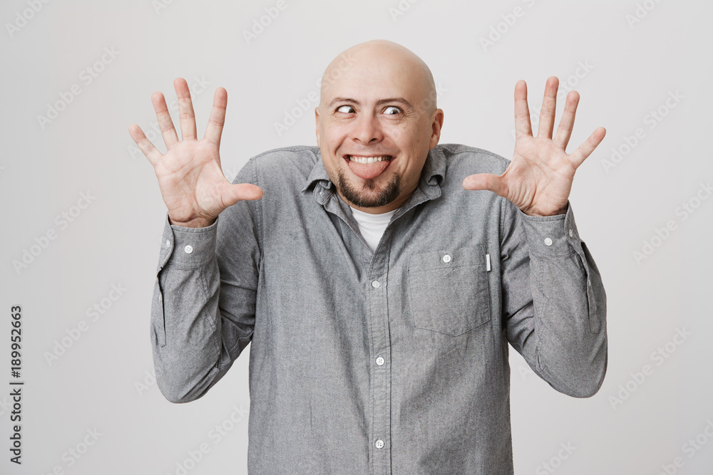 Naklejka premium Indoor shot of bald bearded man making grimace with slanting eyes, posing against gray background with opened palms, sticking out his tongue. Positive face expression and emotions