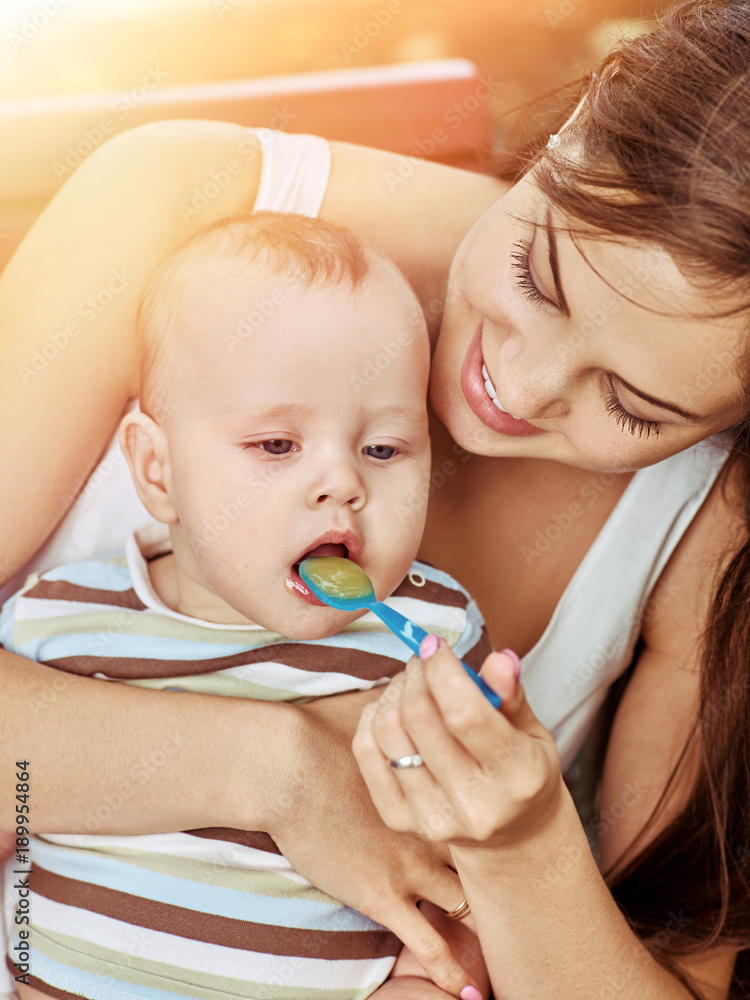 Mother feed her baby porridge by spoon outdoors in park. Spring sunlight of sunset or sunrise. Proper nutrition for children. Toning of the spring photo. Mother feed her baby porridge by spoon outdoors in park. Spring sunlight of sunset or sunrise. Proper nutrition for children. Toning of the spring photo.