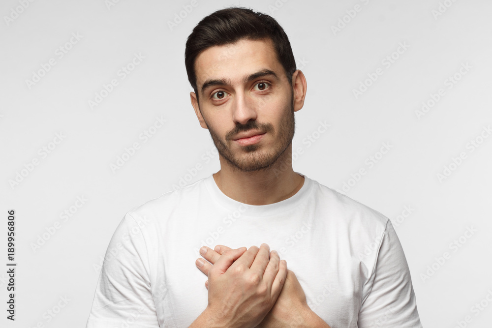 Portrait of smiling positive young man keeps hands on chest, expresses sympathy. Kind hearted friendly nice guy shows kindness, wearing blank white t-shirt Portrait of smiling positive young man keeps hands on chest, expresses sympathy. Kind hearted friendly nice guy shows kindness, wearing blank white t-shirt