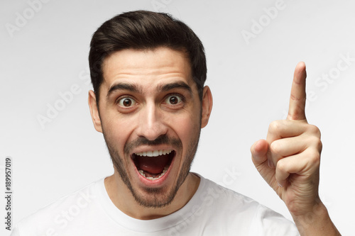 Close up portrait of young handsome smiling man in white t-shirt, pointing his finger in eureka sign, having great innovative idea, understanding or solution he has just got