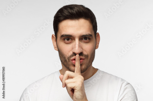 Close up shot of handsome tricky man with shh gesture, asking for silence or to be quiet, isolated on gray background