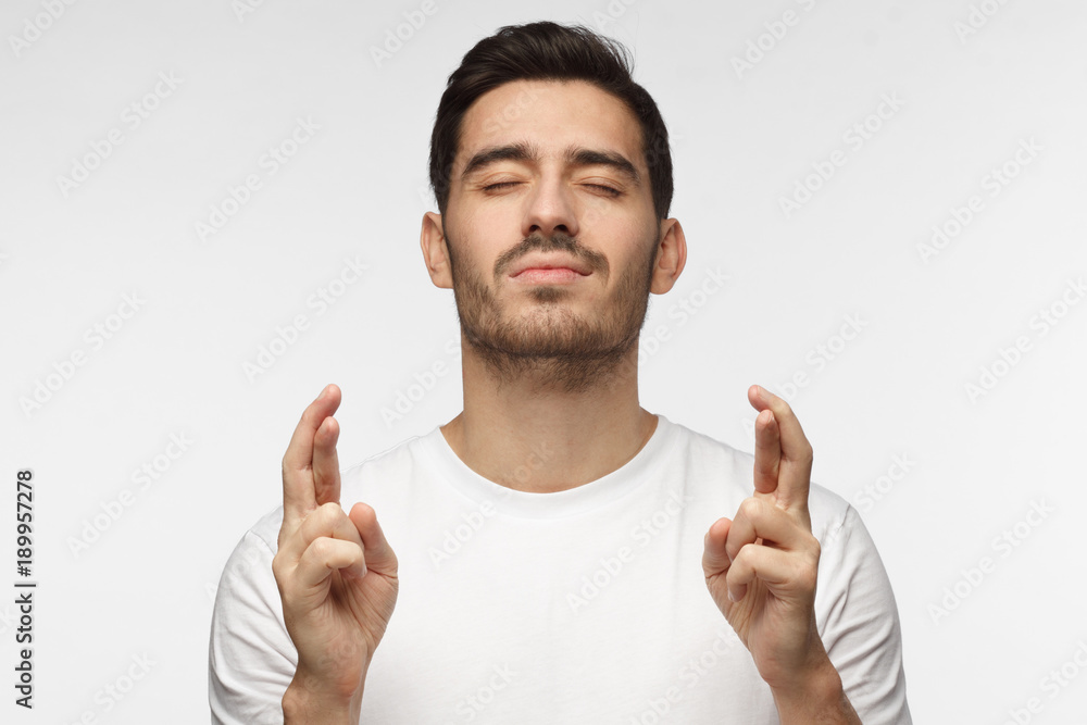 Indoor photo of young man isolated on grey background, looking deeply concentrated with eyes closed and fingers of both hands crossed, wishing luck, praying Indoor photo of young man isolated on grey background, looking deeply concentrated with eyes closed and fingers of both hands crossed, wishing luck, praying