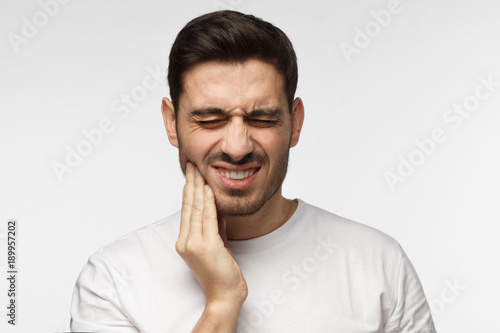 Closeup of young man isolated on gray background touching his face and closing eyes with expression of horrible suffer from health problem and aching tooth, showing dissatisfaction © Damir Khabirov