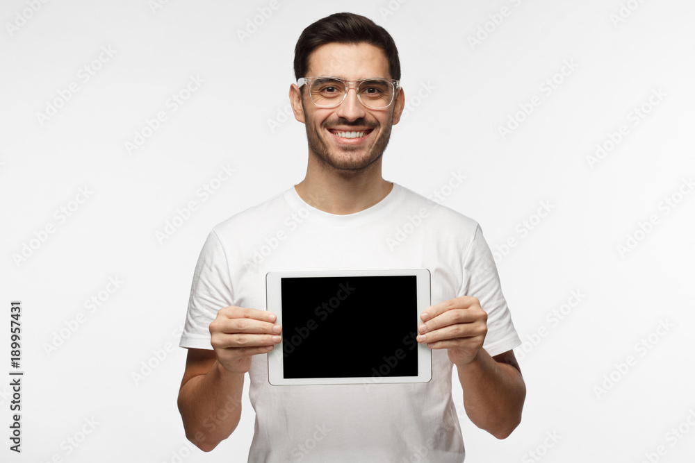 Studio picture of positive man isolated on grey background standing in casual clothes holding tablet and showing it blank screen with happy smile as if advising product or service Studio picture of positive man isolated on grey background standing in casual clothes holding tablet and showing it blank screen with happy smile as if advising product or service