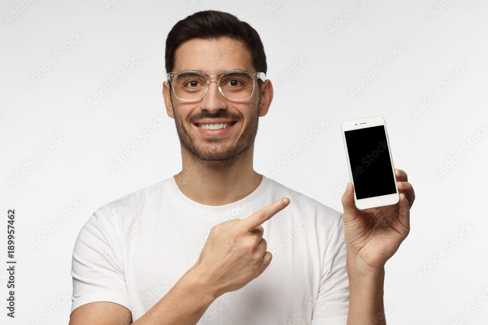 Handsome man in white tshirt in transparent glasses isolated on grey background, presenting smart phone and pointing with finger at blank black screen with copyspace for ads Handsome man in white tshirt in transparent glasses isolated on grey background, presenting smart phone and pointing with finger at blank black screen with copyspace for ads