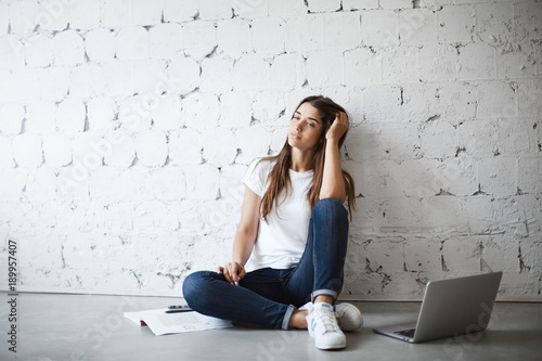 Portrait of dreamy young attractive girl, leaning on wall and sitting on floor with laptop and notes, gazing on something. Cute beautiful teenager thinking about conversation with boy she likes