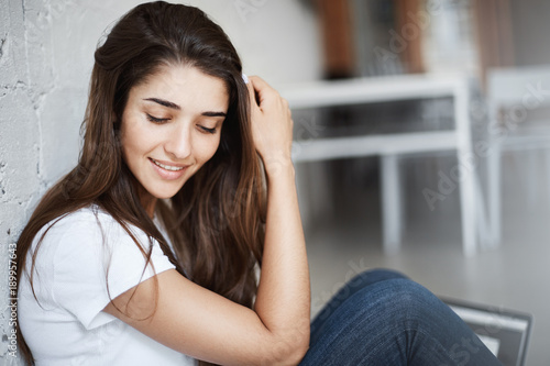 Close-up photo of tender attractive brunette, smiling while touching her hair, leaning on wall and sitting on floor, over blurred background. Shy girlfriend recieves compliments from handsome guy