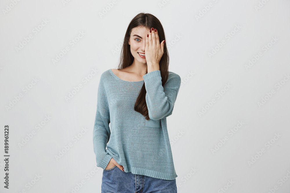 Waist-up portrait of beautiful joyful charming young brunette woman in loose sweater smiling happily, having fun indoors, closing one eye with hand while playing with kids, posing against gray studio Waist-up portrait of beautiful joyful charming young brunette woman in loose sweater smiling happily, having fun indoors, closing one eye with hand while playing with kids, posing against gray studio