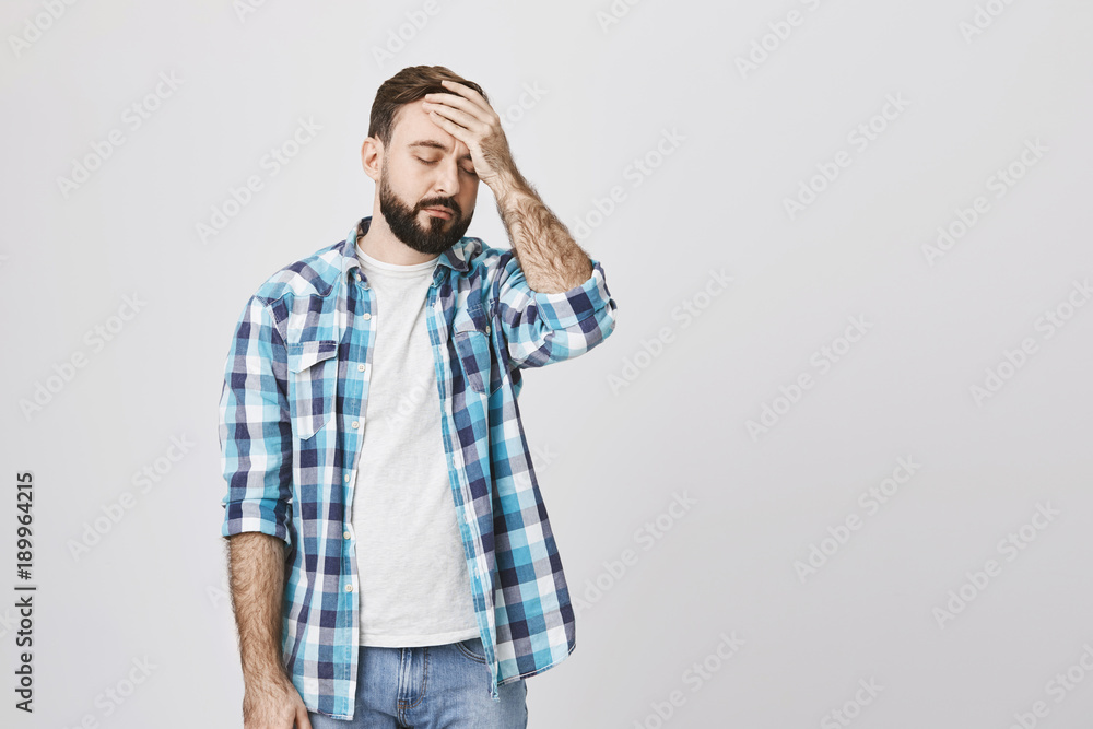Tired unhappy bearded guy in checked shirt, holding hand on forehead, standing against gray background. Male businessman suffers headache because of huge pressure at work Tired unhappy bearded guy in checked shirt, holding hand on forehead, standing against gray background. Male businessman suffers headache because of huge pressure at work