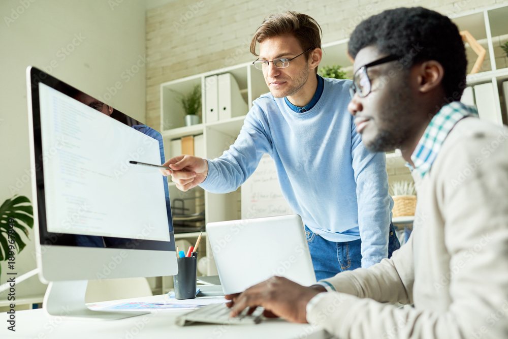 Portrait of two young web developers, one of them African, discussing IT project pointing at code lines on computer screen Portrait of two young web developers, one of them African, discussing IT project pointing at code lines on computer screen