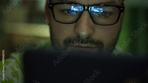 Young man used at smartphone watching a video on digital tablet, the video lights up his face and his glasses reflect the light.