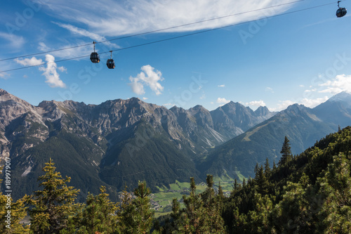 A gondola going up a mountain with a mountain and forest background. The are trees in the foreground and below the gondola. photo
