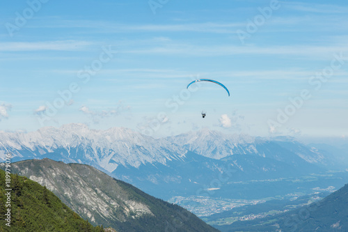 Paraglider flying near a mountain high above towns below. There are mountains in the background Grass is in the foreground. The setting is cool.