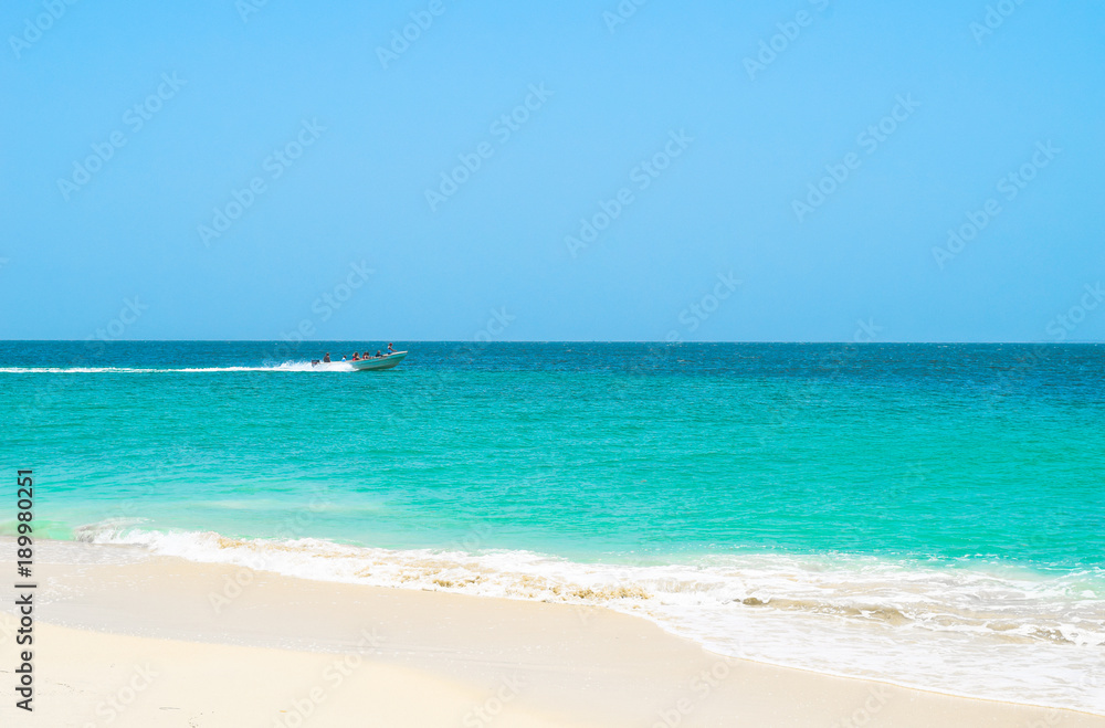 Fototapeta Boot bringt Touristen zur Insel, im Vordergrund weißer Sandstrand, das türkisene Meer und der blaue Himmel, Motorboot im Hintergrund fährt auf Wasser