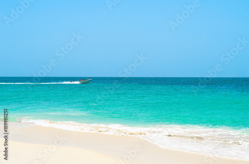 Boot bringt Touristen zur Insel, im Vordergrund weißer Sandstrand, das türkisene Meer und der blaue Himmel, Motorboot im Hintergrund fährt auf Wasser © Laila