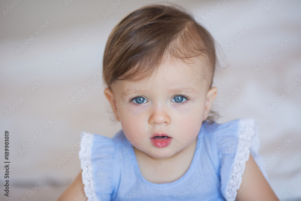 Pretty little baby girl in a blue dress sits on a bed at home