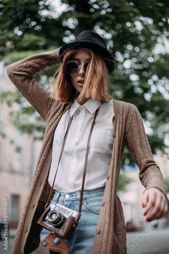 Portrait of fashionable young woman with a camera. Round sunglasses and hat