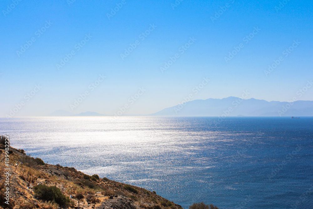 Wunderschöner Ausblick auf das blaue Meer, die Sonne spiegelt sich im Wasser, am Horizont sieht man eine Insel, blauer Himmel