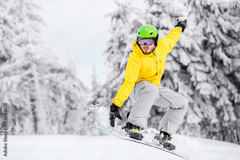 Man in colorful sports clothes jumping with snowboard on the snowy mountains with beautiful trees on the background