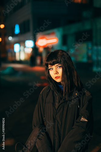 Young Japanese girl stand near neon lights and pose to the camera. Red and blue light, night time, street