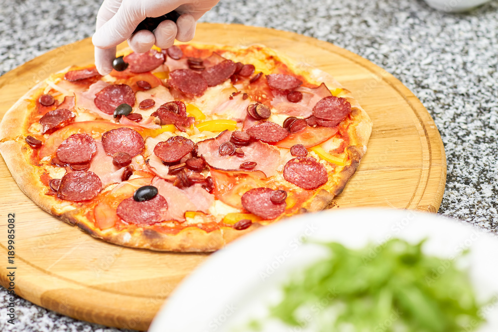 Fototapeta Olives sprinkling on salami pizza. Putting olives onto pizza.