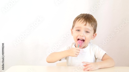 Baby boy smiling and eating lollipops on a stick, sitting at a table on a white background.