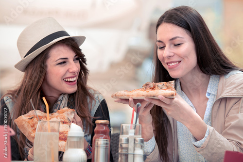 Two smiling girls eating pizza in cafe.