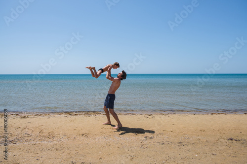 Young happy father holding up in his arms little son putting him up at the beach.