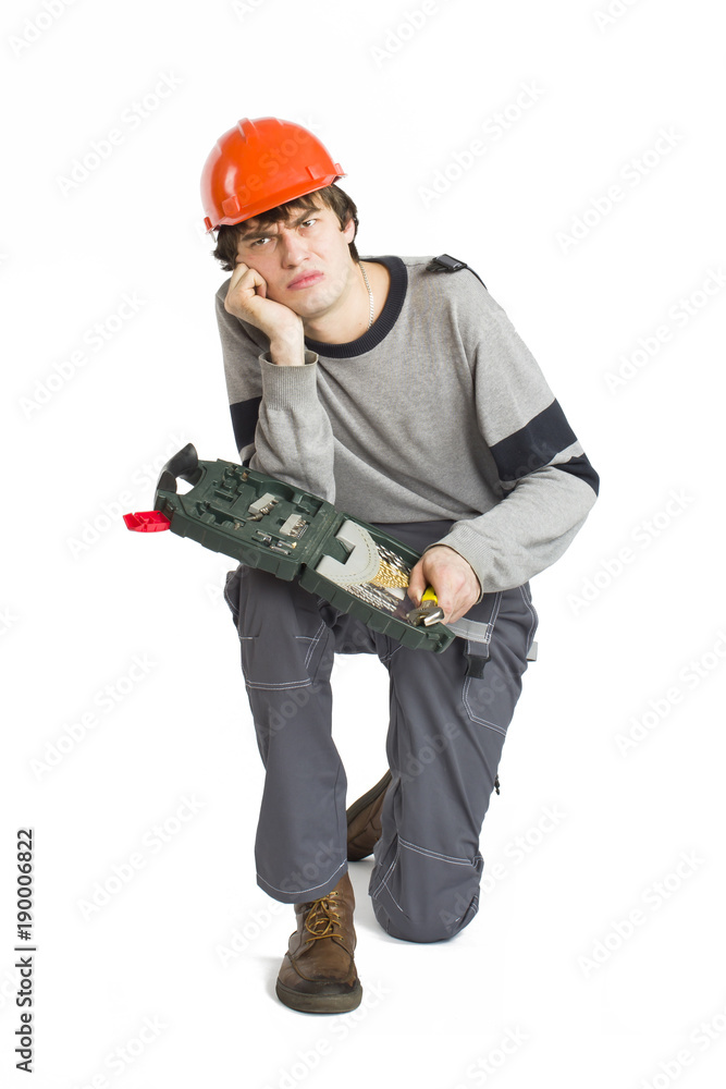 A young man in working grey clothes and orange hard helmet sitting with toolkit on isolated white background. A young man in working grey clothes and orange hard helmet sitting with toolkit on isolated white background.