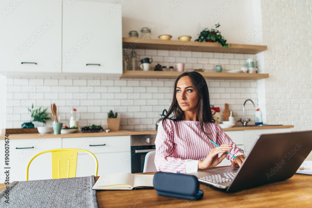 Portrait of young woman sitting at the table in kitchen working on laptop. Portrait of young woman sitting at the table in kitchen working on laptop.