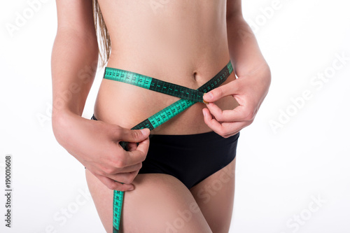 Fit and healthy young lady measuring her waist with a tape measure in centimeters and millimeters. She has her black gym exercise outfit on. Isolated image on white.
