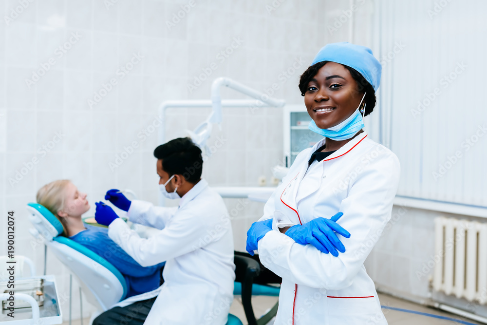 Young smiling African american female dentist in front of dentist checking teeth to patient at clinic. Dental clinic concept. Young smiling African american female dentist in front of dentist checking teeth to patient at clinic. Dental clinic concept.