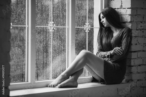 Beautiful young woman sitting alone near window with rain drops. Sexy and sad girl with long slim legs. Concept of loneliness. Black and white.