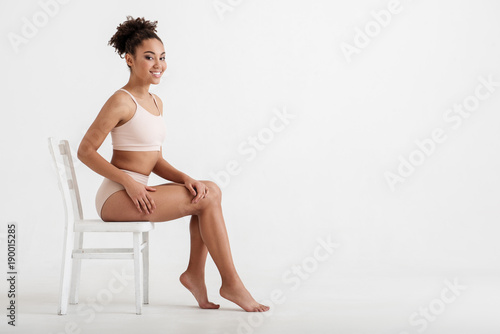 Full length portrait of satisfied girl bragging about her body shape while sitting on stool and smiling. Copy space in right side. Isolated on background