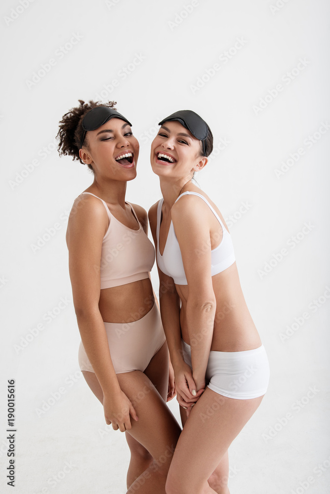 Portrait of slumber laughing girls standing in underwear and winking at  camera. Blindfolds are on their heads. Isolated on background Stock Photo