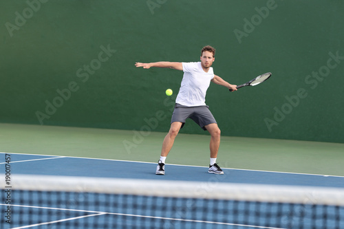 Professional tennis player athlete man focused on hitting ball over net on hard court playing tennis match with someone. Sport game fitness lifestyle person living an active summer lifestyle.