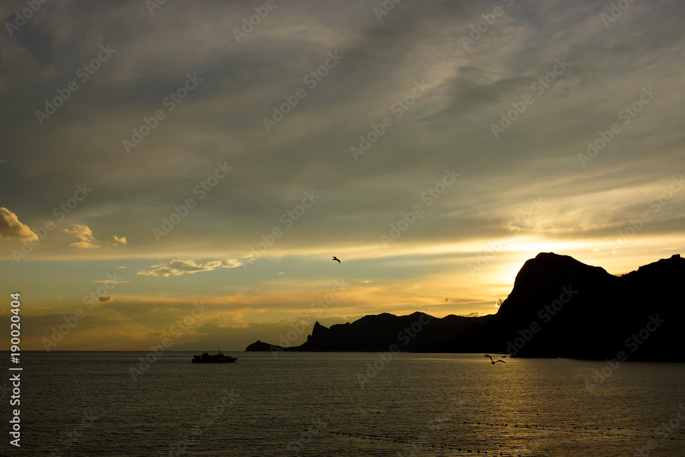 Sunset on the Black Sea. Sudak Bay. The outline of cape Kapchik. A gloomy sunset on the sea and a lonely boat in the bay. Landscapes of the Crimea.
