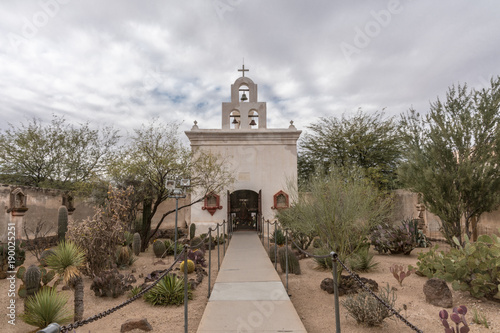 Photo Tucson, Arizona, USA - January 9, 2018: White ide chapel with bell tower for Virgin of Guadalupe at historic San Xavier Del Bac Mission stands in desert garden under heavy gray sky