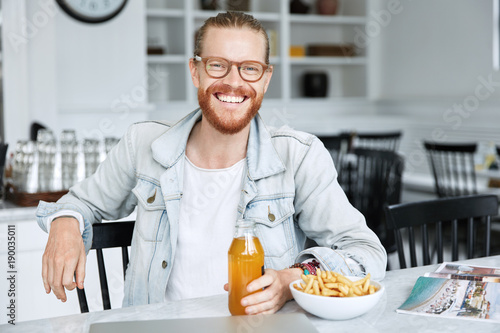 Happy bearded male model in denim jacket  enjoys fresh beverage with french fries  feels relaxed  reads magazine at cafe  happy to meet with friends. People  lunch  leisure and rest concept.
