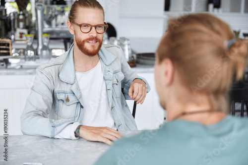 Talented male designer with ginger beard and mustache, dressed in denim jacket, wears spectalces, has meeting with colleague, discuss future project together, share their experience or ideas.