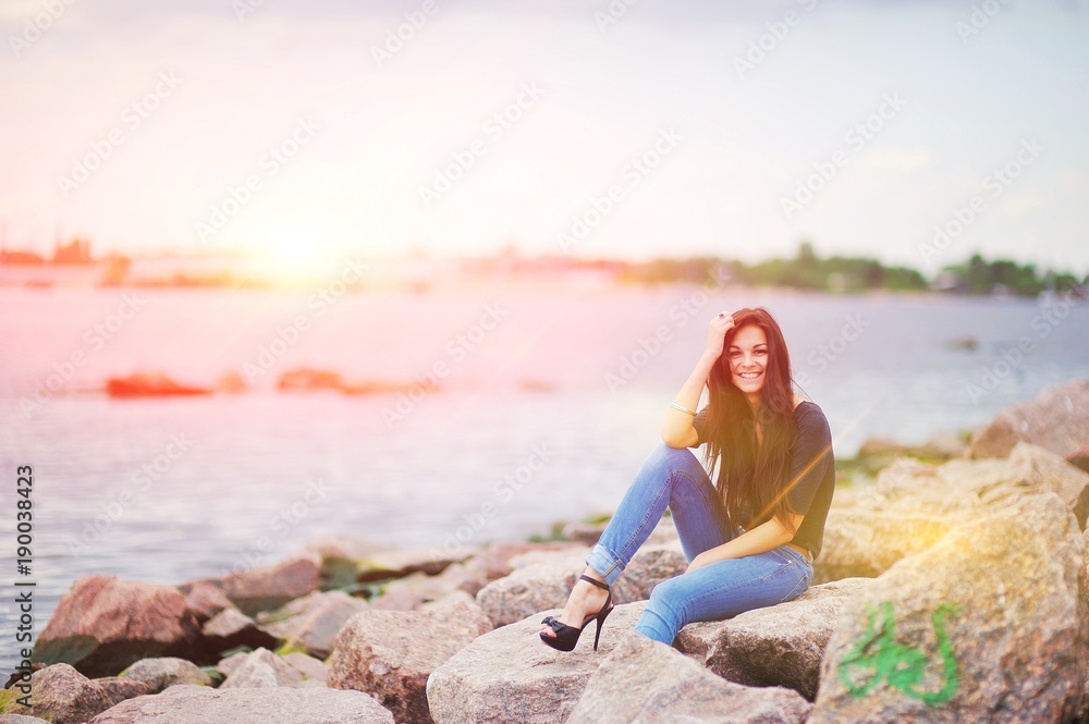 Beautiful smiling girl in black shirt, stylish jeans and shoes on high heels posing while sitting on stones near water.